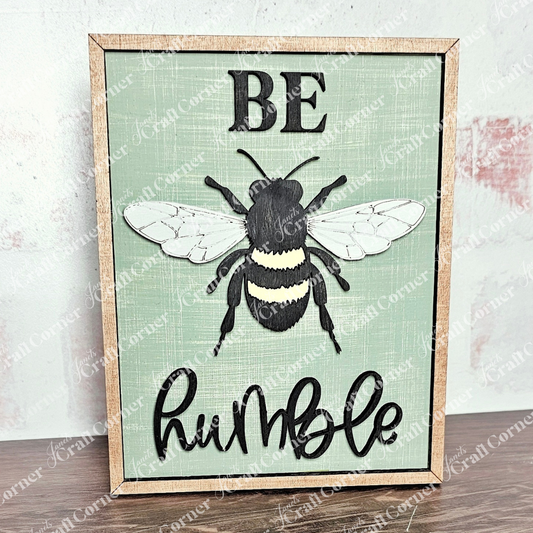 The Be Humble Farmhouse Sign from Janet's Craft Corner showcases a bee illustration on a light green background with "BE" in bold and "humble" in cursive, adding elegance and simplicity to any room's décor.