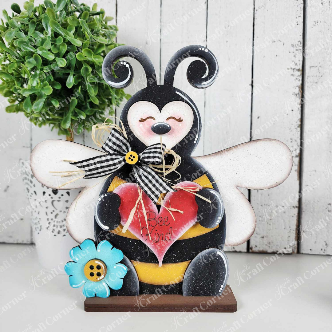 Janet's Craft Corner's Honey Bee Mini Shelf Sitter Set is perfect for bee-lovers, featuring a bee figurine holding a "Bee Kind" heart. Adorned with a bow and blue flower, it complements any wooden background next to a small potted plant.
