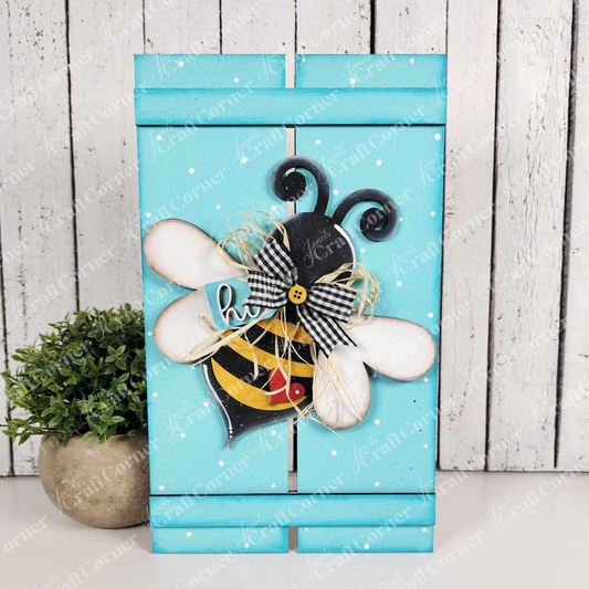 The Bee Pallet Sign Shelf Leaner from Janet's Craft Corner is a turquoise wooden block with a bee cutout featuring white wings, black and yellow stripes, a gingham bow, and a yellow flower button, perfect for home decor or as part of your DIY craft kit collection.