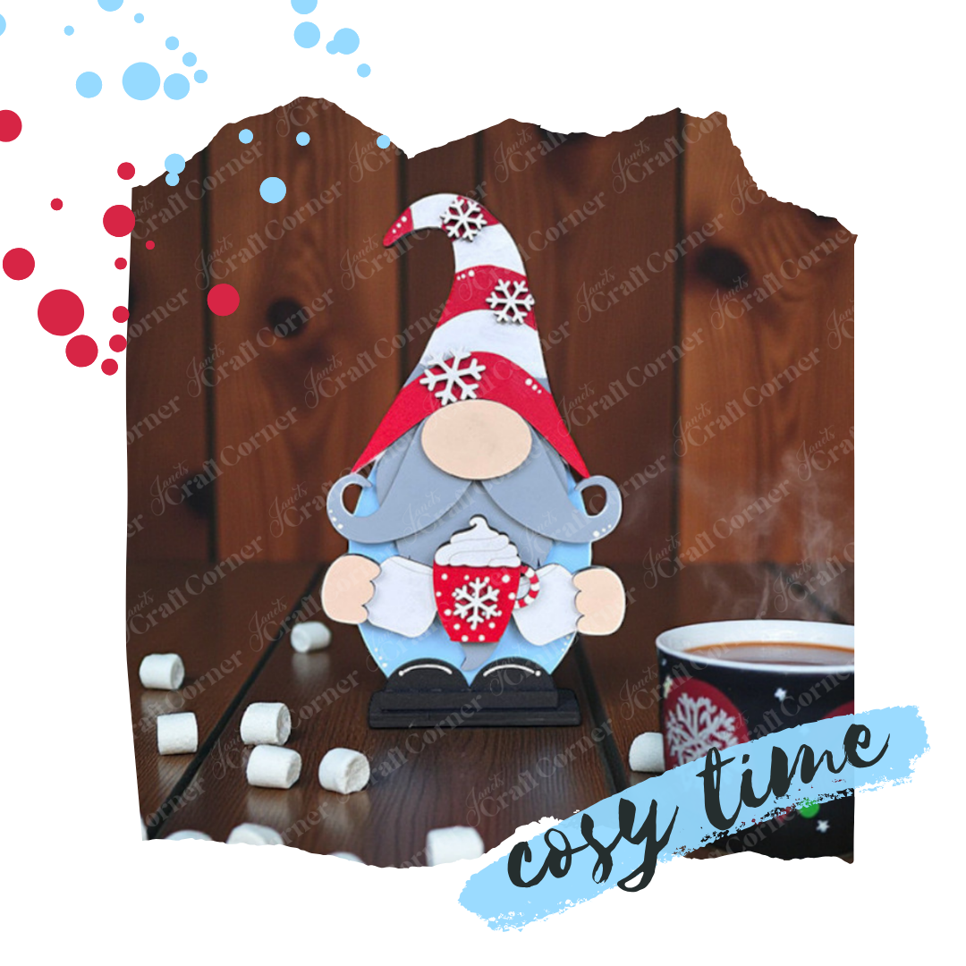 A Hot Cocoa Gnome Shelf Sitter from Janet's Craft Corner stands with a red and white snowflake hat, holding a matching mug on a table with a steaming mug of hot cocoa and scattered mini marshmallows.