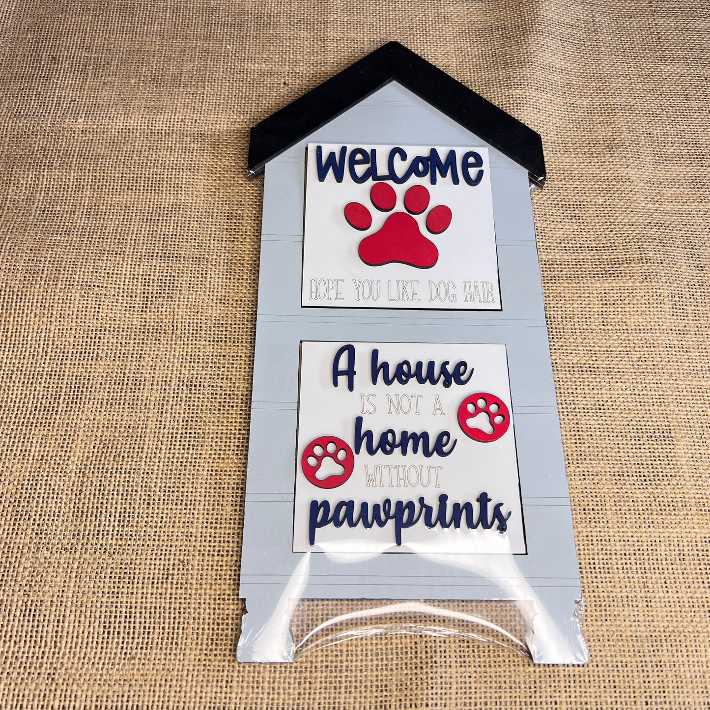 The Heartland House Swappable Squares Holders Shelf Leaner – Painted with Inserts features a doghouse-shaped wood cut-out sign with the message “Welcome, hope you like dog hair. A house is not a home without pawprints,” accented by red paw prints and displayed against a charming burlap background.