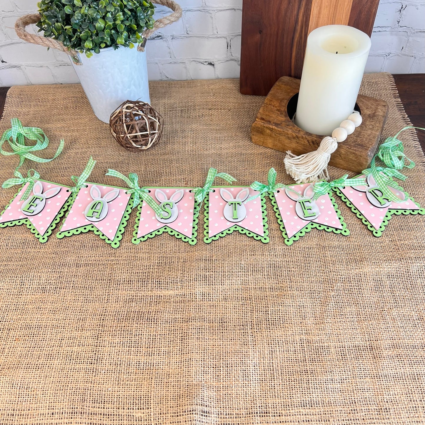 The Easter Pennant Banner from Janet's Craft Corner, featuring pink and green pennants that spell out "EASTER," decorates a burlap-covered table. This charming DIY home décor setup is completed with a potted plant, a candle, and a decorative wooden bead tassel.
