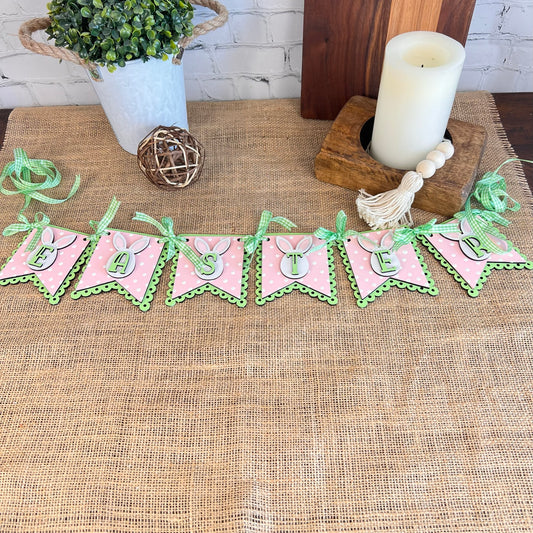 The Easter Pennant Banner from Janet's Craft Corner, featuring pink and green pennants that spell out "EASTER," decorates a burlap-covered table. This charming DIY home décor setup is completed with a potted plant, a candle, and a decorative wooden bead tassel.