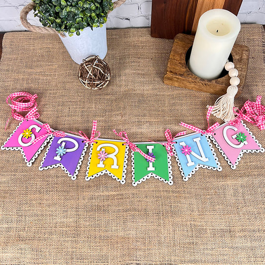 A vibrant "SPRING" banner from Janet's Craft Corner brings life to the burlap surface, featuring pink checkered ribbons and floral details enhancing its charm. Complementing this home décor piece, you'll find a potted plant, a woven ball, a wooden block with a white candle on top, and a tassel—ideal elements for your DIY Spring Banner kit.