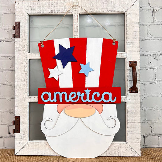 This door decoration by Janet's Craft Corner, the Uncle Sam Gnome Door Hanger, features a patriotic cartoon face with a white beard and mustache. It sports a red and white striped hat adorned with blue stars and the word "America," making it perfect for an Independence Day display on a rustic white door with glass panels.