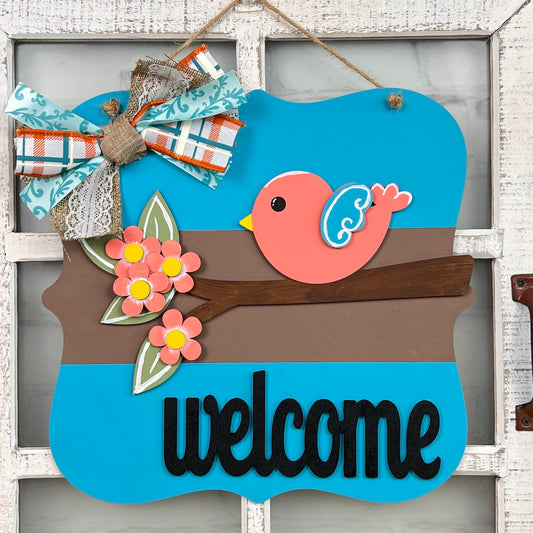The Bird Welcome Sign by Janet's Craft Corner features a vibrant pink bird with blue wings perched on a brown branch, highlighted by delicate pink flowers. A stylish bow interweaving floral and plaid designs adds a decorative touch to the corner. Ideal for home décor, it elegantly displays the word "welcome" in black at the bottom.
