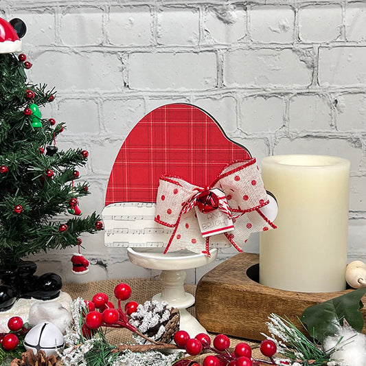 A festive setup showcasing the Chunky Santa Hat from Janet's Craft Corner, adorned with a red plaid pattern, white base, and bow, elegantly perched on a stand. This centerpiece is complemented by holiday-themed ornaments, a glowing candle, evergreen branches, and red berries set against a white brick wall background—ideal for enhancing your holiday home decor.