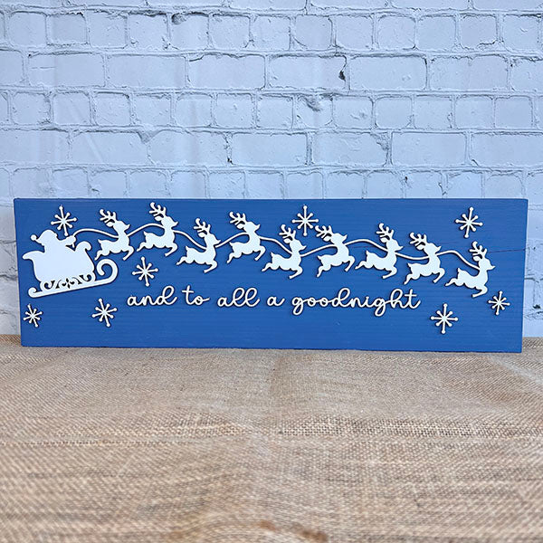 The Santa Sleigh Mantle Shelf Sitter by Janet's Craft Corner is a blue wooden sign showing Santa’s sleigh and reindeer with snowflakes in white, and "and to all a goodnight" in cursive, displayed against a brick wall backdrop.