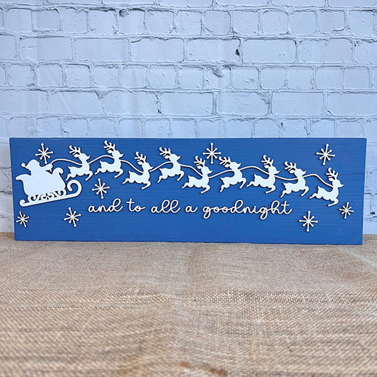 The Santa Sleigh Mantle Shelf Sitter by Janet's Craft Corner is a blue wooden sign showing Santa’s sleigh and reindeer with snowflakes in white, and "and to all a goodnight" in cursive, displayed against a brick wall backdrop.