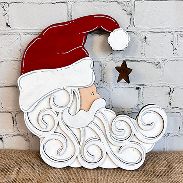 The Santa Moon Shelf Leaner by Janet's Craft Corner is a charming decorative wooden figure featuring a red hat and a white, curly beard, ideal for your Christmas decor. Hanging from the tip of the hat is a small star inscribed with "2022," perfect against a white brick wall. Enhance any space with this delightful piece, available as both a DIY Kit or ready-made option.