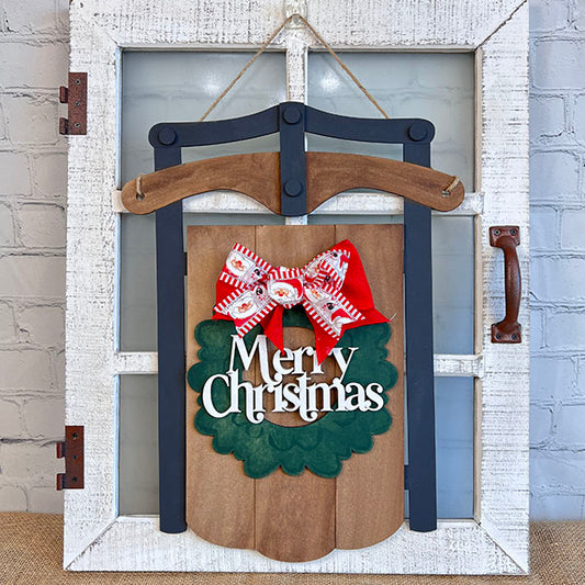 A Merry Christmas Sled by Janet's Craft Corner is beautifully displayed on a rustic white window frame, featuring a green felt design with "Merry Christmas" text. It's enhanced with a charming red and white bow, making it a perfect addition to your home décor against the brick wall.