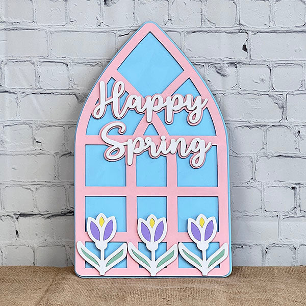 This charming piece of home décor from Janet's Craft Corner, the Spring Tulips Window, is a decorative wooden plaque shaped like an arched window. It features "Happy Spring" in pink cursive with three tulip designs at the bottom, set against a light blue background and a textured white brick wall.