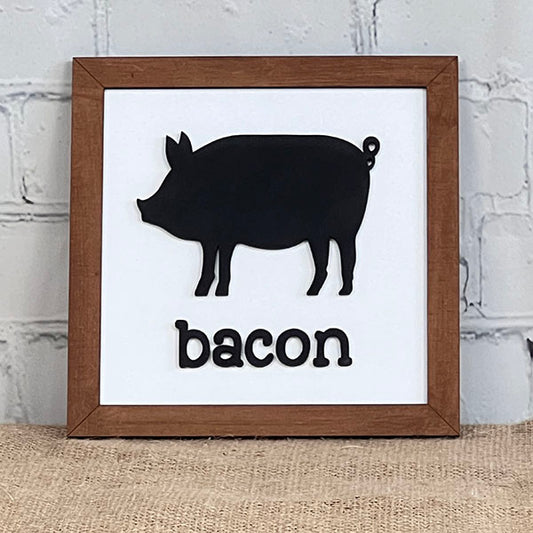 The "Bacon Eggs Milk Signs" from Janet's Craft Corner, ideal for kitchen decor, showcases a black silhouette of a pig positioned above the word "bacon." The wooden frame borders the image against a white brick wall and rests on a burlap surface.