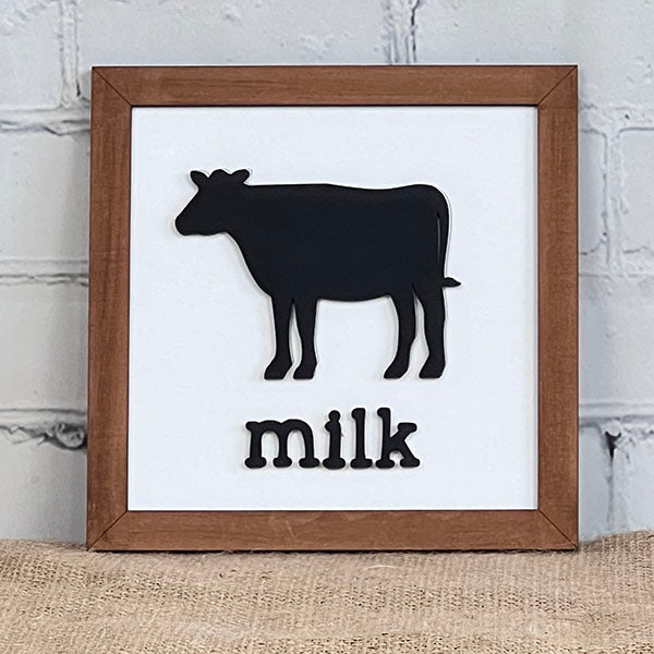 The "Bacon Eggs Milk Signs" by Janet's Craft Corner showcase a framed artwork with a black silhouette of a cow above the word "milk" in black text, making it charming kitchen decor. The frame is placed on a burlap surface against a white brick wall backdrop, adding rustic flair to any culinary space.