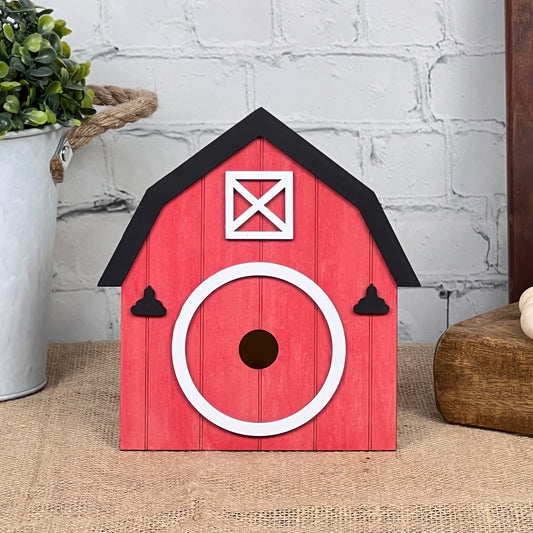 The Barn Interchangeable Set by Janet's Craft Corner is a decorative red birdhouse styled like a barn. It features a round white entrance and black roof accents, set against a brick wall backdrop. This charming piece of home décor is beautifully complemented by a potted plant and wooden block, infusing any space with rustic charm.