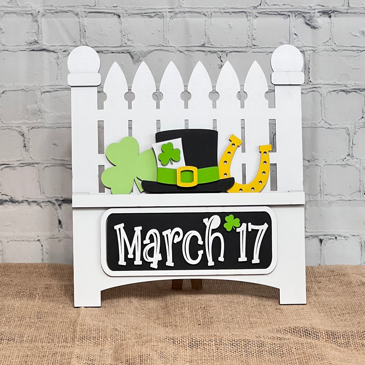 A decorative white fence display against a brick wall features interchangeable inserts from Janet's Craft Corner celebrating St. Patrick's Day, including a green shamrock, a black hat with a shamrock, and a yellow horseshoe. The plaque reads "March 17" with a small shamrock accent.