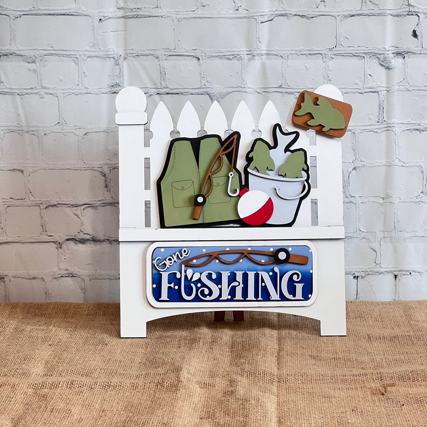 The Gone Fishing Interchangeable Insert craft kit by Janet's Craft Corner includes a DIY wood picket fence with fishing gear, a vest, bobber, fish bucket, and fish sign. A blue "Gone Fishing" sign sits at the bottom; perfect for seasonal home decor.