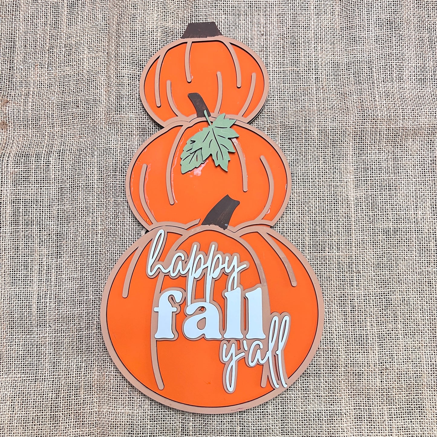 The Reversible Snowman/Pumpkin Shelf Leaner by Janet's Craft Corner is a DIY seasonal home decor kit featuring three stacked orange pumpkins on burlap; the bottom pumpkin reads "happy fall y'all," and a green leaf accents the center.