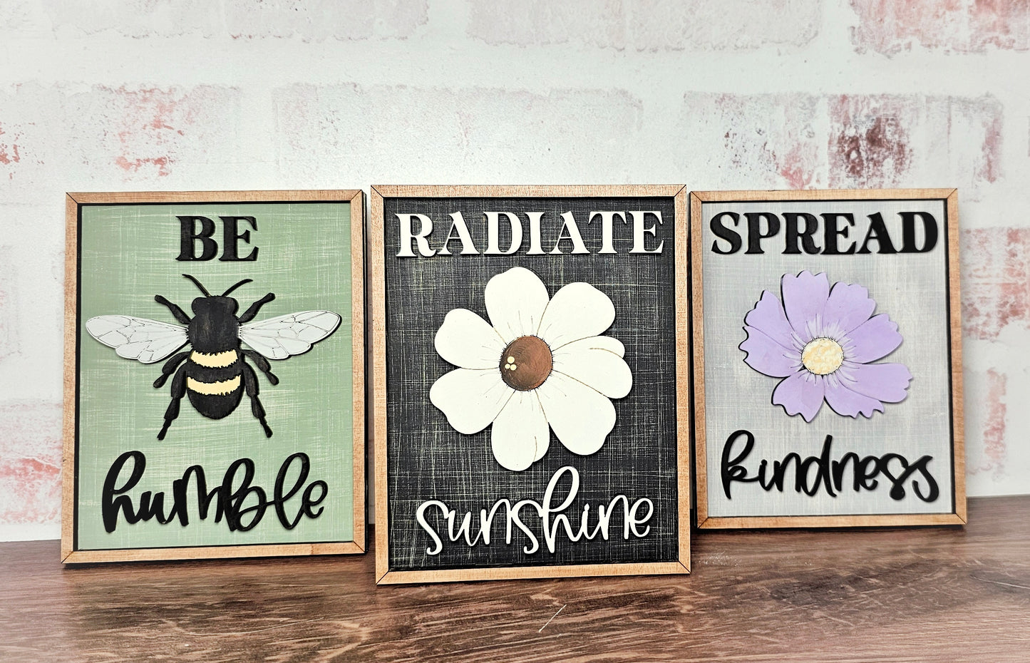 Three signs from Janet's Craft Corner show motivational messages: "Be Humble" with a bee, "Radiate Sunshine" with a white flower, and "Spread Kindness" with a purple flower. Displayed on wood against a light brick background, they're ideal as farmhouse home décor.
