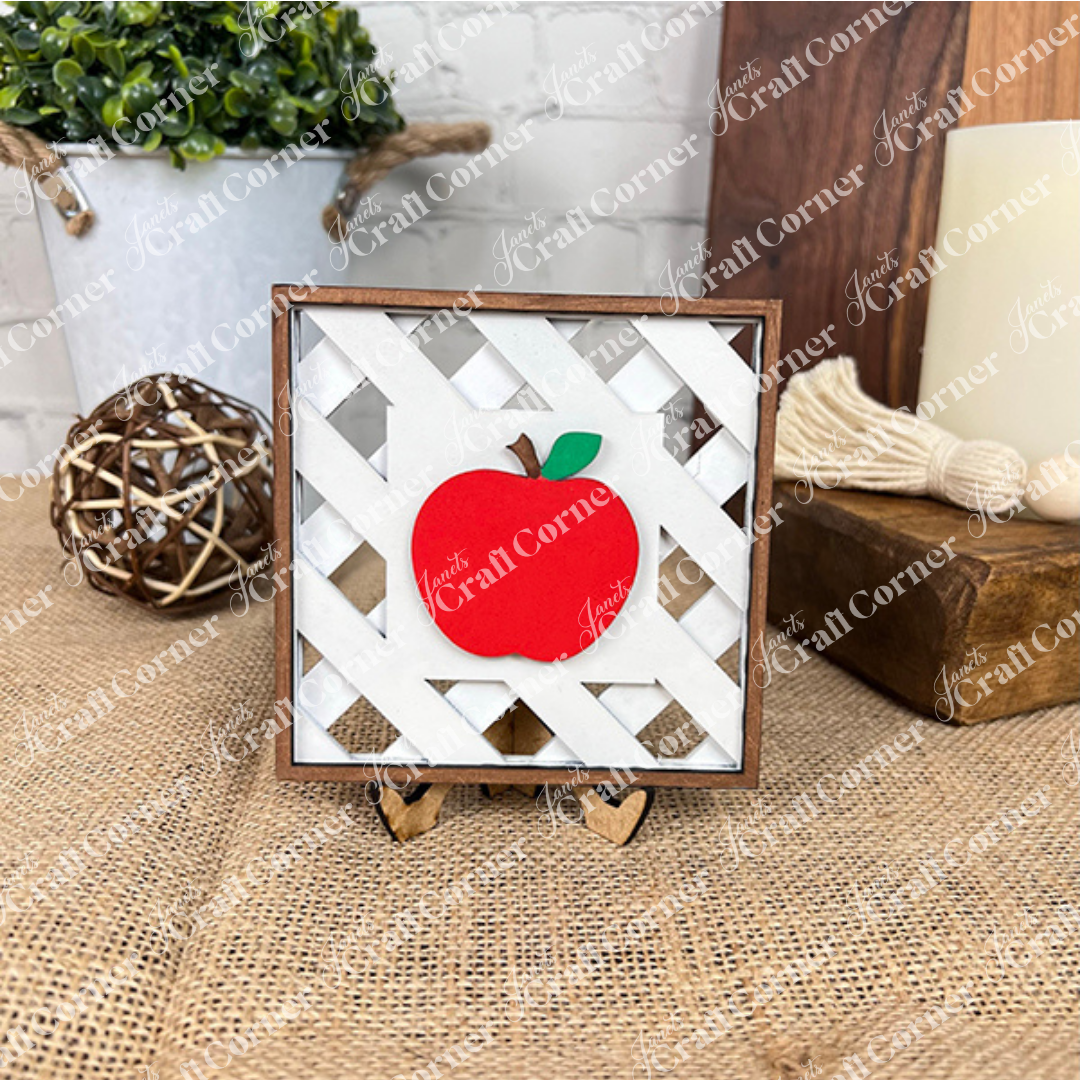 Janet's Craft Corner's Apple Swappable Squares DIY kit features a red apple wooden cut-out with a green leaf on a white lattice square frame and easel—ideal for rustic home decor and seasonal craft lovers. Set of 1.