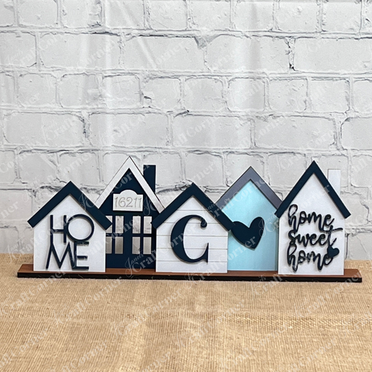 Showcase the Family Houses Shelf Sitter by Janet’s Craft Corner—a DIY home decor craft kit featuring five charming house shapes, each with unique designs and heartfelt words such as “HOME” and “home sweet home.” Beautifully arranged in front of a white brick wall, this set makes for perfect personalized décor or a delightful family houses shelf sitter.