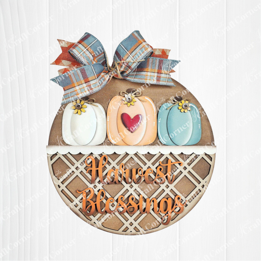 The Harvest Blessings Wall Hanger by Janet's Craft Corner is a DIY seasonal decor kit featuring a round sign with white, blue, and orange pumpkins topped with yellow flowers, a plaid bow, and "Harvest Blessings" in orange letters. Set of 1.