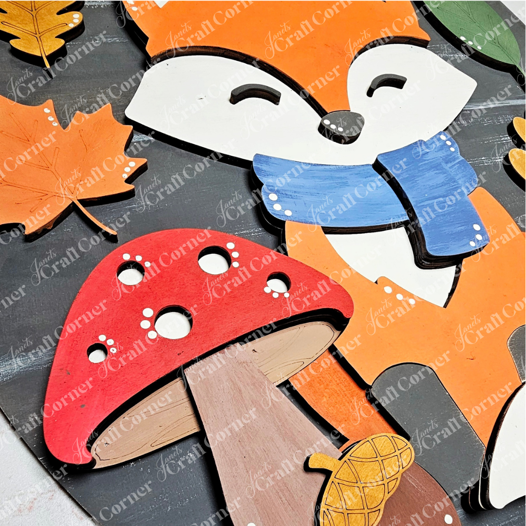 The Hello Fox Oval Door Hanger by Janet's Craft Corner is a DIY seasonal craft kit featuring a smiling fox in a blue scarf, an orange leaf, mushroom, and acorn—perfect for adding woodland charm to your home.