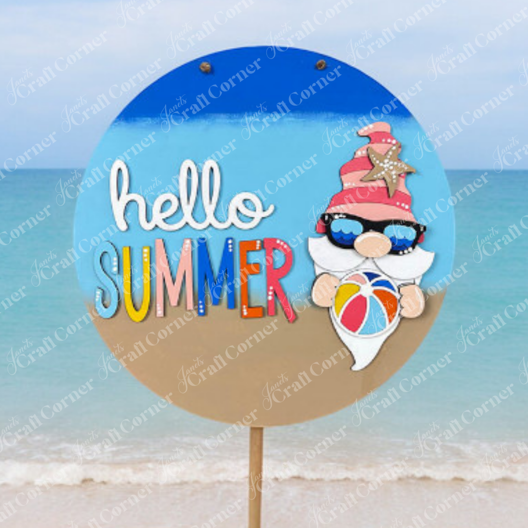 A round sign on a beach reads "hello SUMMER" in colorful letters, featuring Janet's Craft Corner’s 10.5" Hello Summer Gnome Wall Hanger DIY Kit with a gnome in sunglasses and starfish hat against an ocean backdrop.