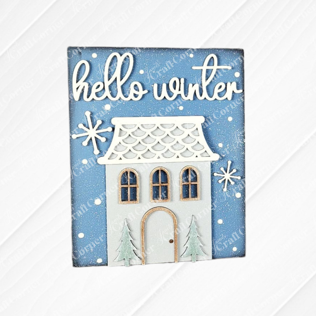 Janet's Craft Corner Welcome Winter Switchable Farmhouse Signs—DIY seasonal home decor kit featuring a house, pine trees, snowflakes, and “hello winter” in cursive—perfect for cozy winter decorating. Painted with a blue sky and white house it makes a cute addition to any shelf.