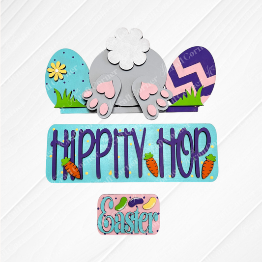The Hippity Hop Bunny Butt Interchangeable Insert from Janet's Craft Corner is a charming DIY craft kit featuring a bunny tail, colorful eggs, paw prints, and "Hippity Hop" in blue with carrot accents on a white wooden background.