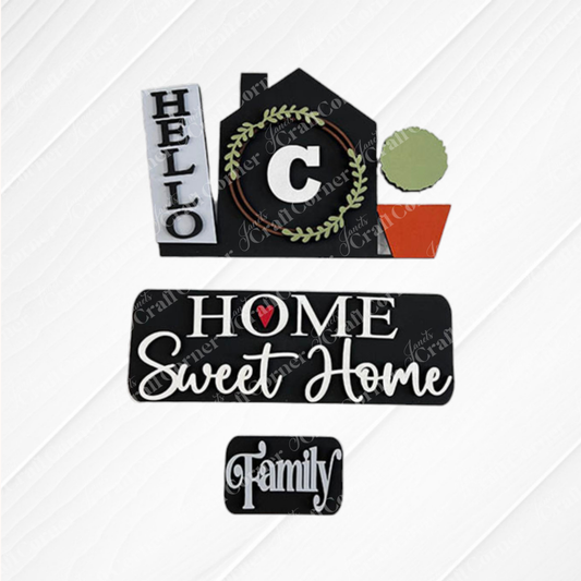 The "Hello Home Monogram Interchangeable Insert" from Janet's Craft Corner is a decorative sign featuring the phrases "Hello," "Home Sweet Home," and "Family." It is designed in the shape of a house with a letter "C," complemented by a green wreath and a small potted plant on a textured white background, making it an ideal piece for versatile home decor.