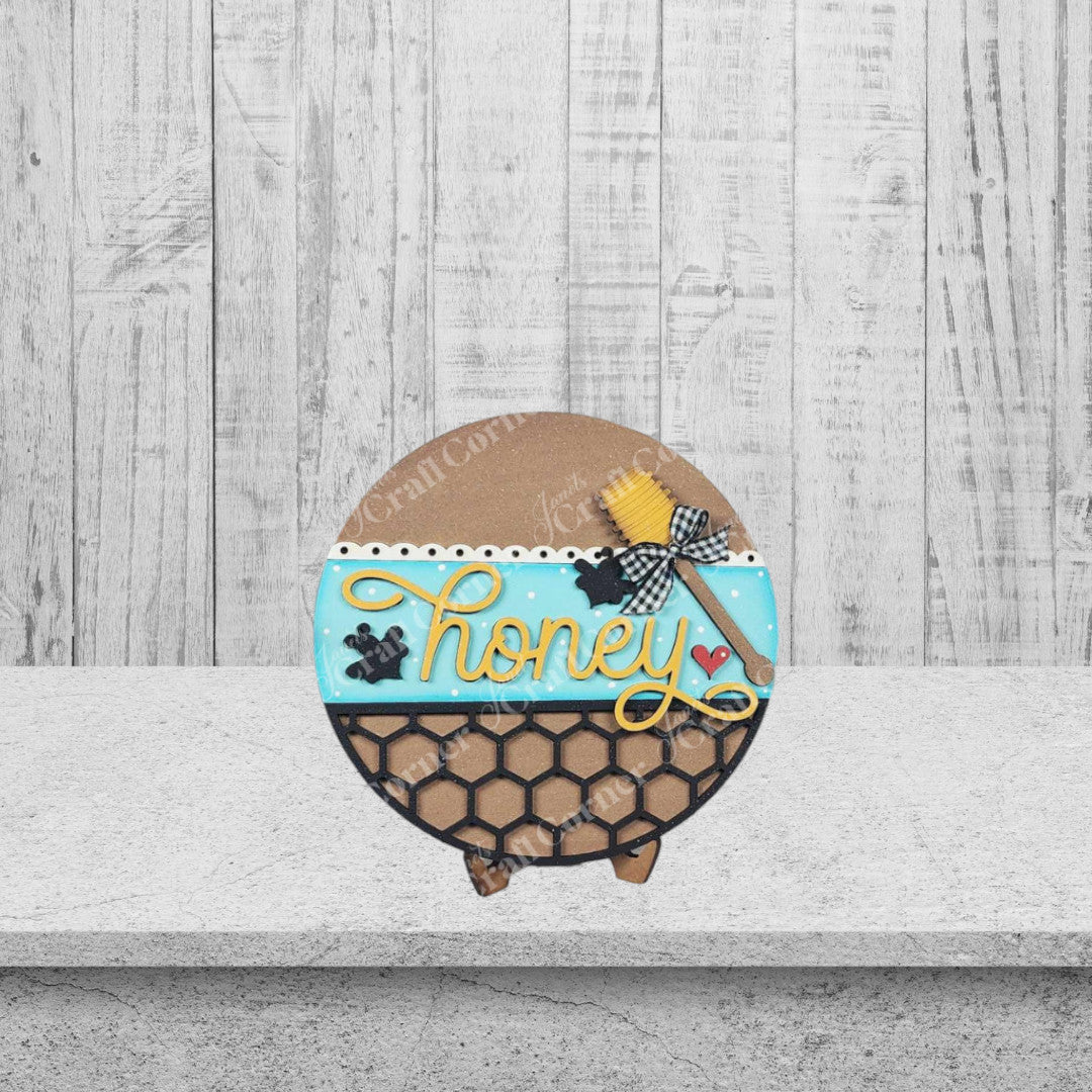 Inspired by Janet's Craft Corner Honey Bee Mini Shelf Sitters DIY kit, a round cookie with blue icing, black honeycomb, yellow "honey," a bee, and red heart is perfect for creating your own seasonal home decor craft.