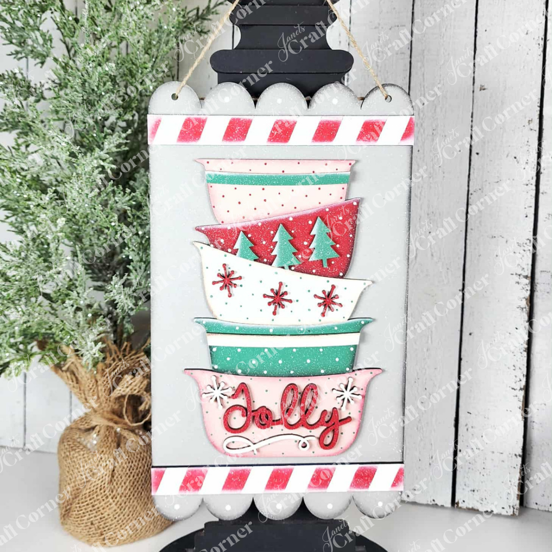 The Jolly Bowls Door Hanger from Janet's Craft Corner is a DIY seasonal home decor kit featuring stacked vintage bowls with festive tree and snowflake patterns, red and white striped edges, and "Jolly" on the bottom bowl. Set of 1.