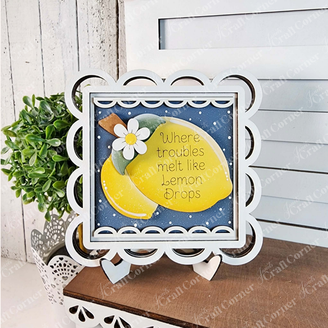Janet’s Craft Corner’s Scallop Lace Square Frame features two lemons, a white flower, and “Where troubles melt like lemon drops” on blue—perfect DIY seasonal decor for your home. Shown on an easel by a white wall and potted plant.