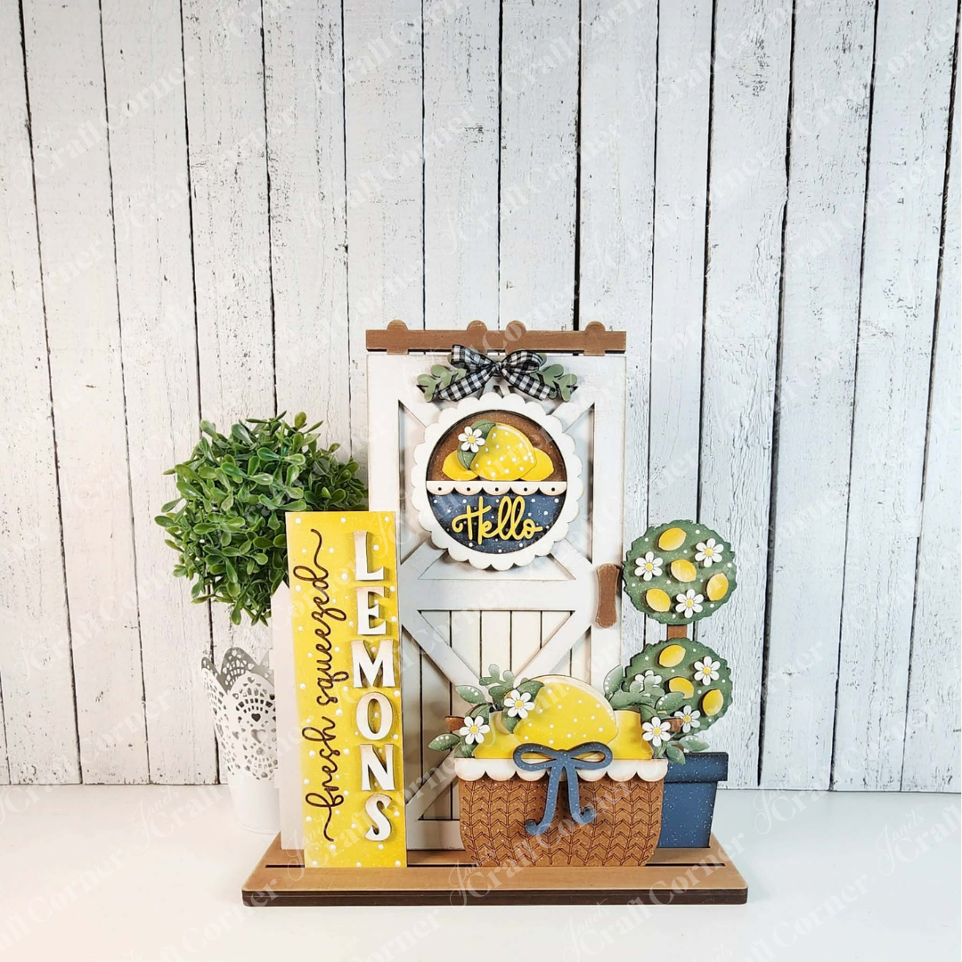 Janet's Craft Corner Lemons Interchangeable Porch Set features a potted green plant, yellow "Lemons" sign, lemon and daisy-themed ornaments, and customizable DIY signs—perfect for farmhouse-style decor on a rustic white wooden wall.