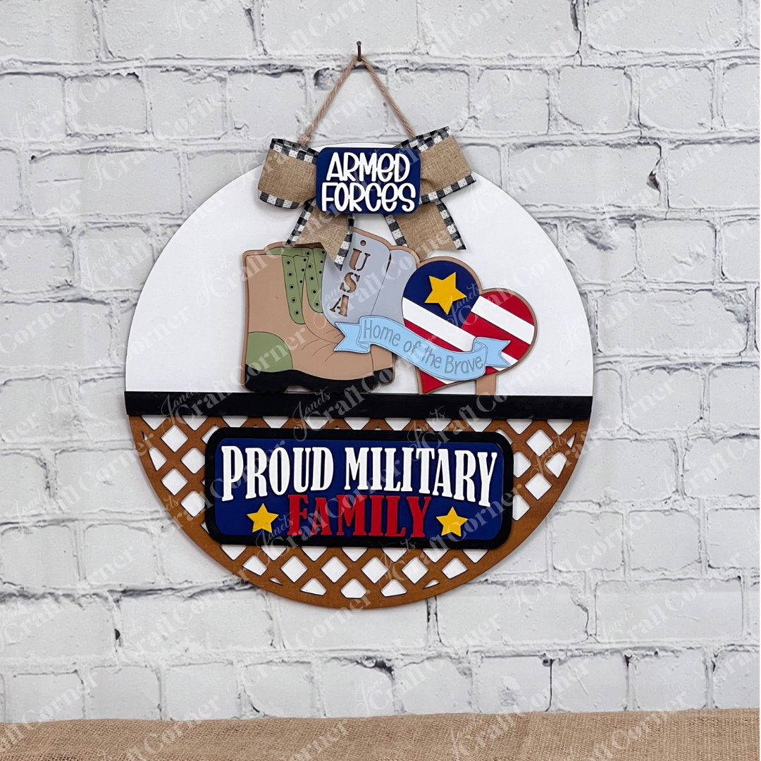A DIY home decor kit by Janet's Craft Corner includes a circular insert featuring a boot, USA tag, stars and stripes with a heart, an "Armed Forces" plaque, and "Proud Military Family" at the bottom. It is accented with a black and white bow on a brick background.