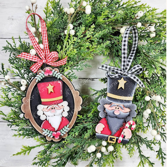 Two Nutcracker ornaments from Janet’s Craft Corner, each with checked ribbon, hang on a green wreath with white berries. Both feature tall red hats topped with gold stars and hold candy canes—one ornament is framed in an oval while the other stands freestanding. These DIY holiday craft kit ornaments are a charming choice for your Christmas tree or as the perfect complement to your favorite festive projects.