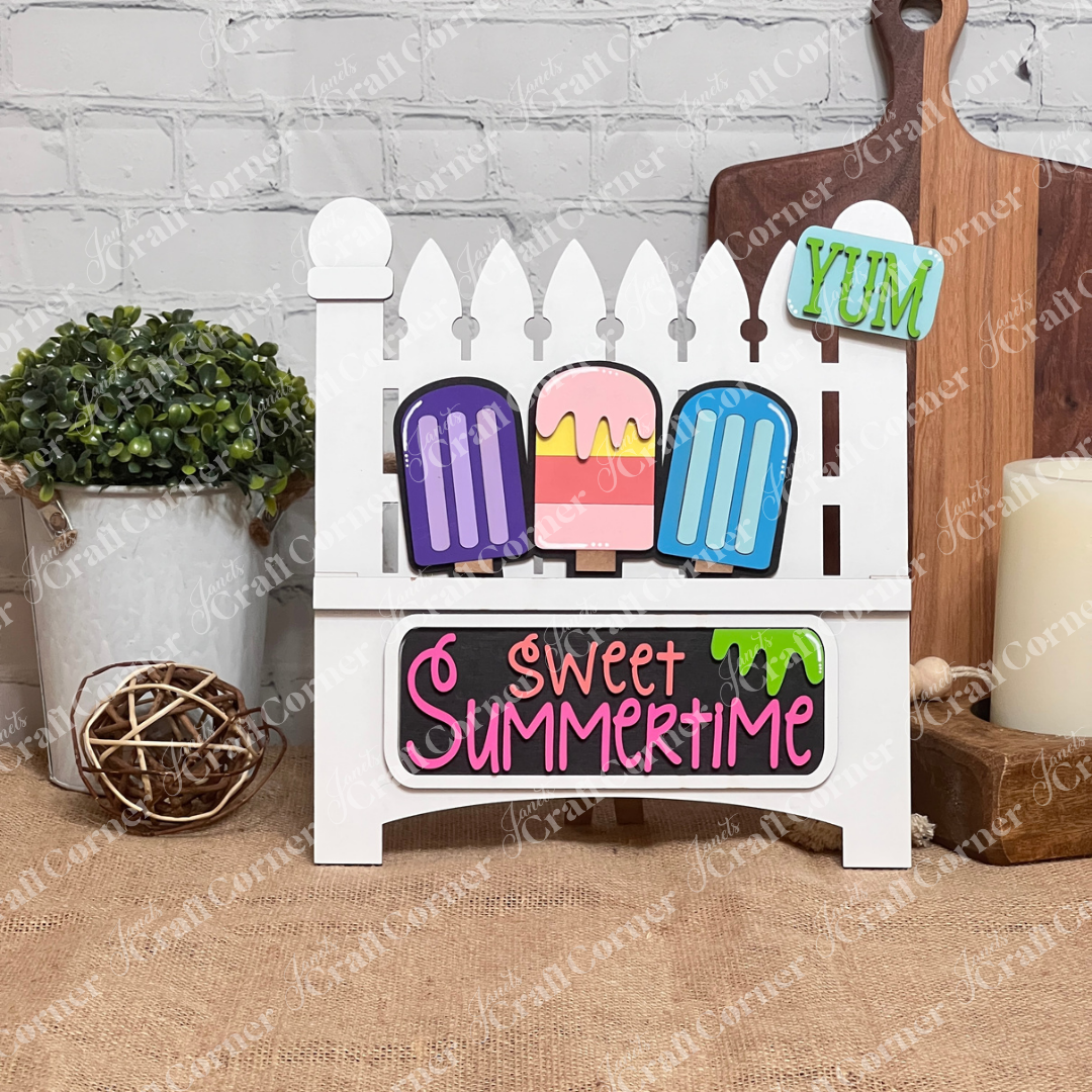 Add a charming touch to your home décor with Janet's Craft Corner's DIY home decor kit, featuring the Popsicle Interchangeable Insert. This decorative piece showcases a white garden fence with vibrant popsicles and "Sweet Summertime" elegantly written in pink and orange above a teal "YUM" license plate. It’s perfect for placing next to a potted plant on a wooden surface.