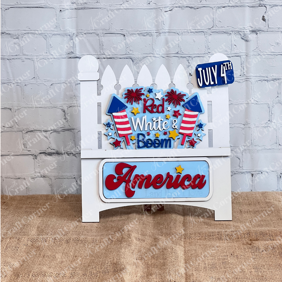 Janet's Craft Corner offers a "Red White & Boom Interchangeable Insert" featuring a patriotic white garden fence with fireworks and the phrases "Red White & Boom" and "America Land that I Love," plus a "July 4th" license plate, all on a white brick background—perfect for DIY seasonal home decor.