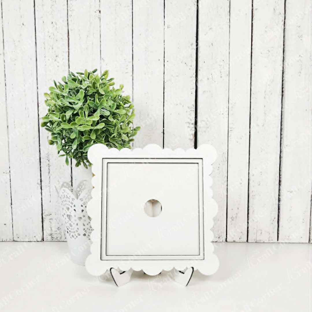 A small green potted plant in a white lace container sits beside Janet's Craft Corner's Scallop Ruffle Square Frame, ideal for swapping wall art and DIY seasonal decor, all set against a white wooden plank background.