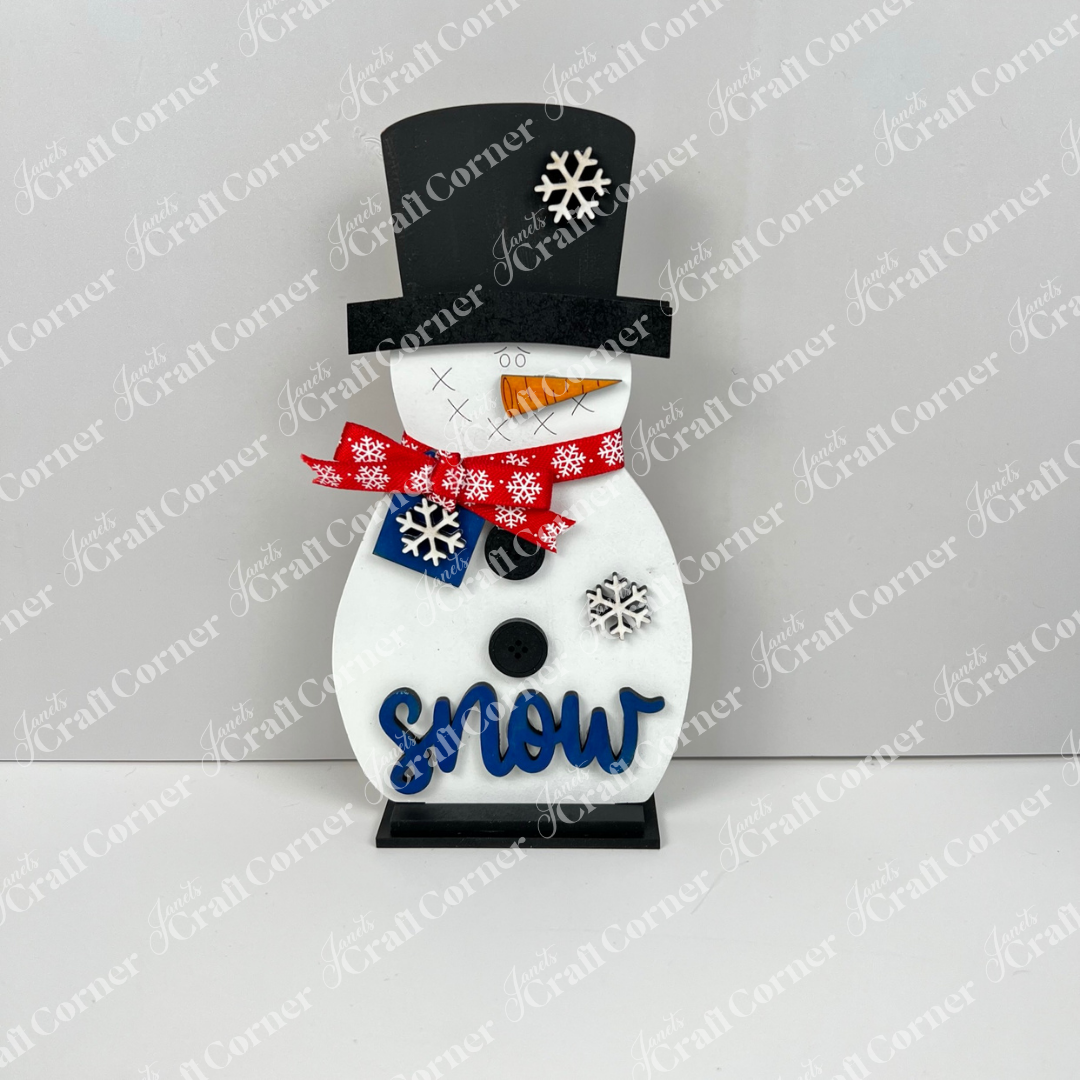 Janet's Craft Corner Snowman Shelf Sitters feature a black top hat, red snowflake scarf, carrot nose, blue "snow" text, and two white snowflake ornaments—perfect festive or winter home decor.