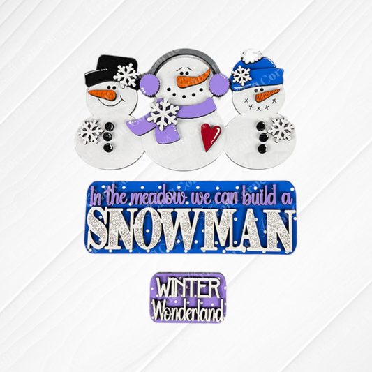 Using the Snowman Interchangeable Insert DIY home decor kit from Janet's Craft Corner, three snowmen adorned with hats and scarves stand against a decorative snowy background. A sign reading "In the meadow, we can build a SNOWMAN" is depicted in purple and blue letters, while below it, a smaller sign declares "WINTER Wonderland." This scene is beautifully accented with snowflakes for an interchangeable home decor feel.
