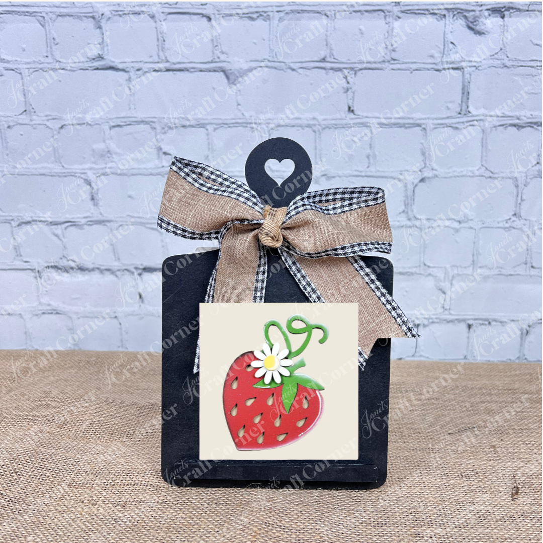 A small black chalkboard with a heart cutout and ribbon bow displays the Strawberry Swappable Squares DIY craft kit by Janet's Craft Corner—featuring a strawberry with a white flower, perfect for seasonal decor accents.