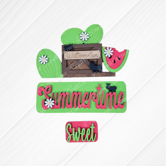 Decorative pieces from Janet's Craft Corner are arranged on a textured white square with a burlap background. The Summertime Watermelon Interchangeable Insert features a wooden watermelon sign alongside green and red "Summertime" and "Sweet" cutouts, small daisies, and hand-painted black elephant shapes for the perfect summer flair.
