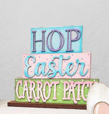 The Easter Bunny Decor Set - Painted by Janet’s Craft Corner features three colorful wooden blocks stacked with the words “HOP,” “Easter,” and “CARROT PATCH,” creating a charming farmhouse-style accent, while a small portion of a white bunny ear peeks out from the corner.