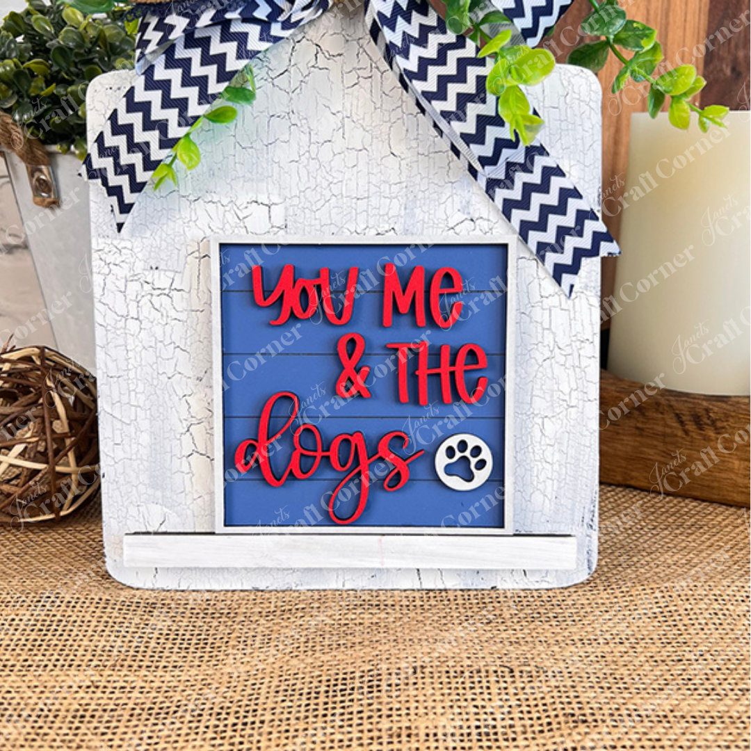 The Dog Squares sign by Janet's Craft Corner features a rustic white frame, blue background, red "You Me & The dogs" text with a paw print, chevron bow, faux greenery, and sits on a woven mat—ideal for tiered tray decor.