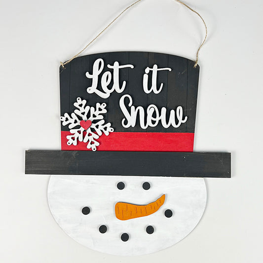The Snowman Door Hanger from Janet's Craft Corner showcases a black top hat with a red band and intricate white snowflake details. Featuring the phrase "Let it Snow" written in elegant cursive, its delightful face includes a carrot-shaped nose and coal-inspired eyes. It's an ideal addition for holiday home décor fans who enjoy whimsical DIY kits.
