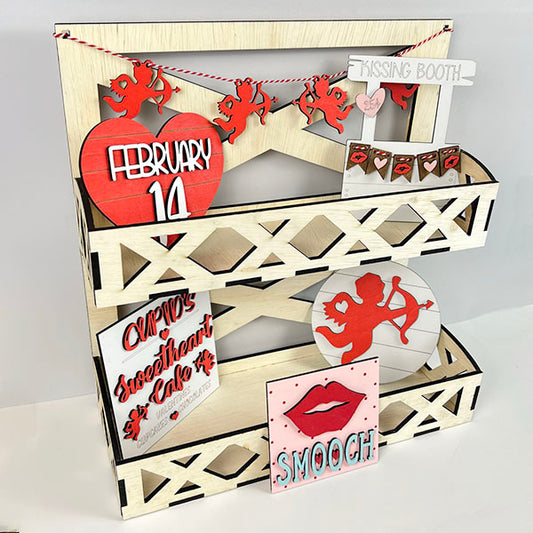 A charming display from Janet's Craft Corner's Cupid Kissing Booth Tiered Tray Kit showcases delightful Valentine's Day decor, featuring a "February 14" heart, cherub cutouts, a playful "Cupid Kissing Booth" sign, and a whimsical "Smooch" sign with lips. The tiered tray boasts a geometric cut-out design ideal for any DIY kit enthusiast.