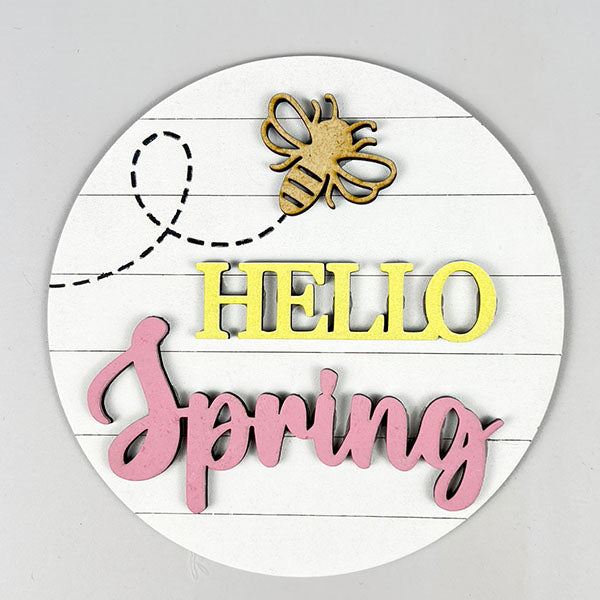 Part of Janet's Craft Corner's Spring Truck Tiered Tray Kit, this charming spring decor features a round sign embellished with a wooden bee cutout. The greeting "HELLO Spring" is creatively presented, with "HELLO" in vibrant yellow block letters and "Spring" in graceful pink cursive against a grooved white backdrop.
