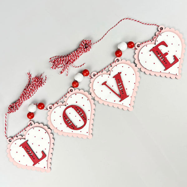Product Description: The L-O-V-E Banner from Janet's Craft Corner is a DIY kit that includes a decorative garland featuring four heart-shaped pieces, each displaying a letter to spell "LOVE." These hearts are elegantly designed with a white finish and pink edges, embellished with red and white beads. They are connected by a stylish red and white twisted cord, making it an ideal addition to your home décor.